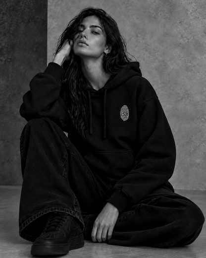 Le Hoodie Signature noir porté par une femme assise — empreinte M.E brodée sur la poitrine