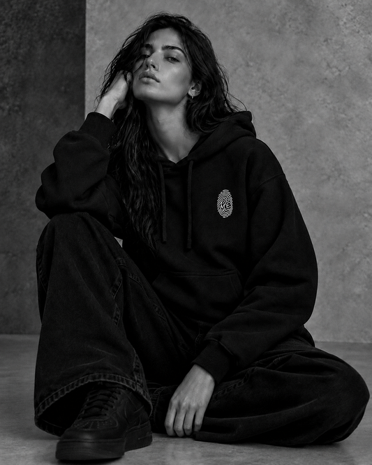 Le Hoodie Signature noir porté par une femme assise — empreinte M.E brodée sur la poitrine